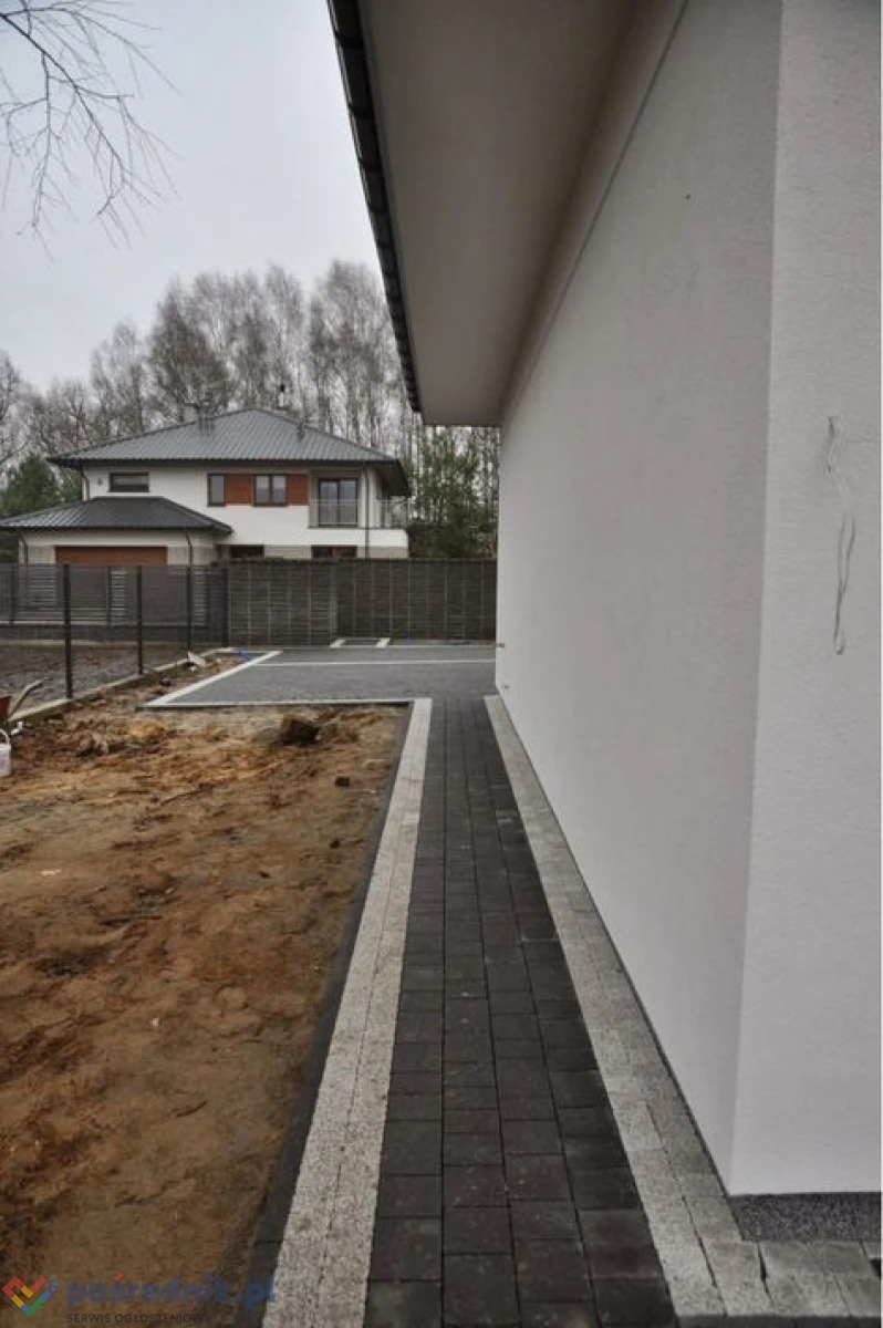 Nowoczesny dom jednorodzinny z jasną elewacją i nowo ułożoną kostką brukową w kolorze grafitowym z jasnymi obrzeżami, widok z boku, w tle drugi dom i drzewa.