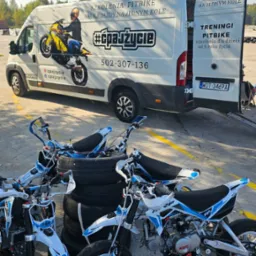 Biała furgonetka z grafiką pitbike i numerem telefonu, zaparkowana obok stosu opon i kilku pitbike'ów Kayo w kolorach białym i niebieskim.