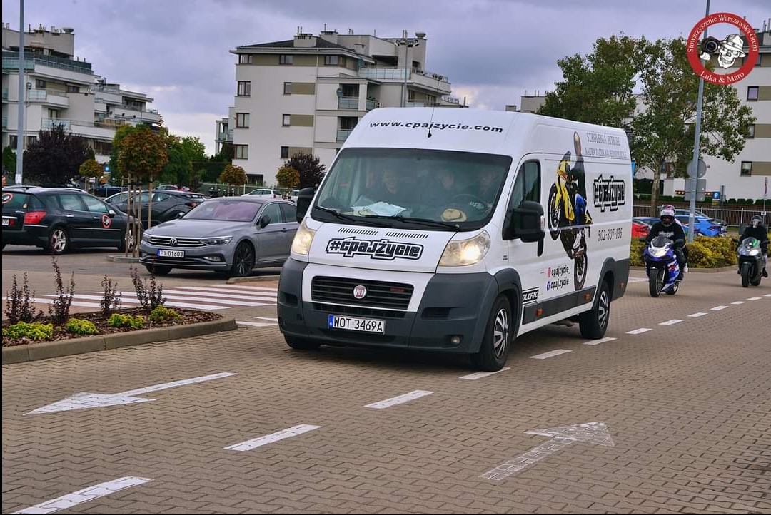 Biały bus z reklamą szkoły jazdy motocyklowej na tle miejskiego krajobrazu, z dwoma motocyklistami jadącymi za nim.