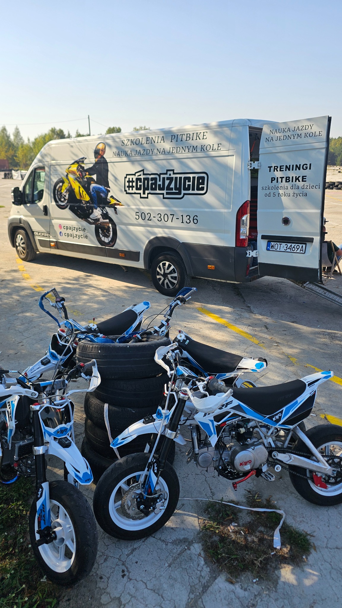 Biała furgonetka z grafiką pitbike i numerem telefonu, zaparkowana obok stosu opon i kilku pitbike'ów Kayo w kolorach białym i niebieskim.