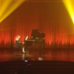 Art-Live Sp. z o.o. - Scena teatralna z pianistką grającą na fortepianie, oświetlona dwoma reflektorami tworzącymi żółte stożki światła na czerwonym tle kurtyny.