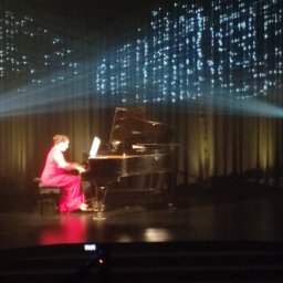 Art-Live Sp. z o.o. - Pianistka w czerwonej sukni gra na fortepianie na scenie z oświetleniem punktowym i projekcją świetlną w tle.