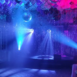 Art-Live Sp. z o.o. - Sala udekorowana na imprezę z balonami pod sufitem, oświetleniem punktowym i wiszącymi serpentynami, widoczna scena i osoba stojąca z boku.