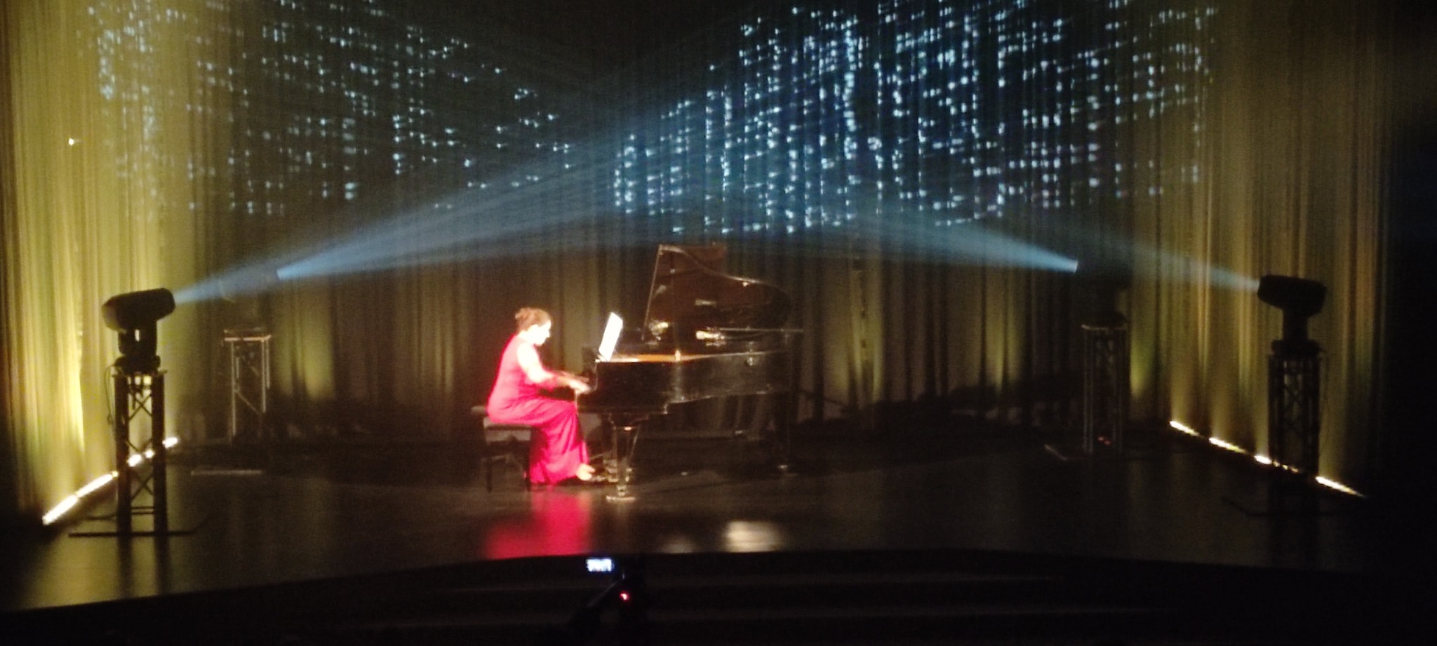 Pianistka w czerwonej sukni gra na fortepianie na scenie z oświetleniem punktowym i projekcją świetlną w tle.