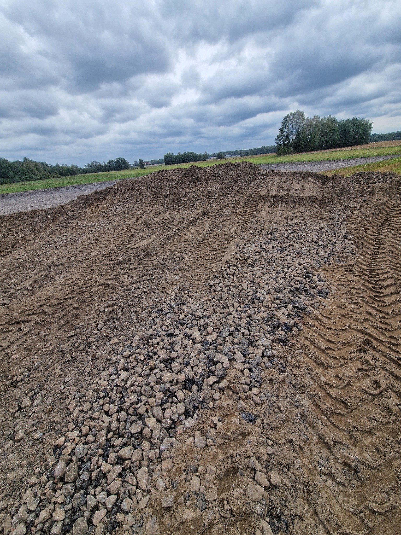 Przygotowany teren pod budowę z widocznymi śladami opon i częściowo wysypanym żwirem, pochmurne niebo w tle.