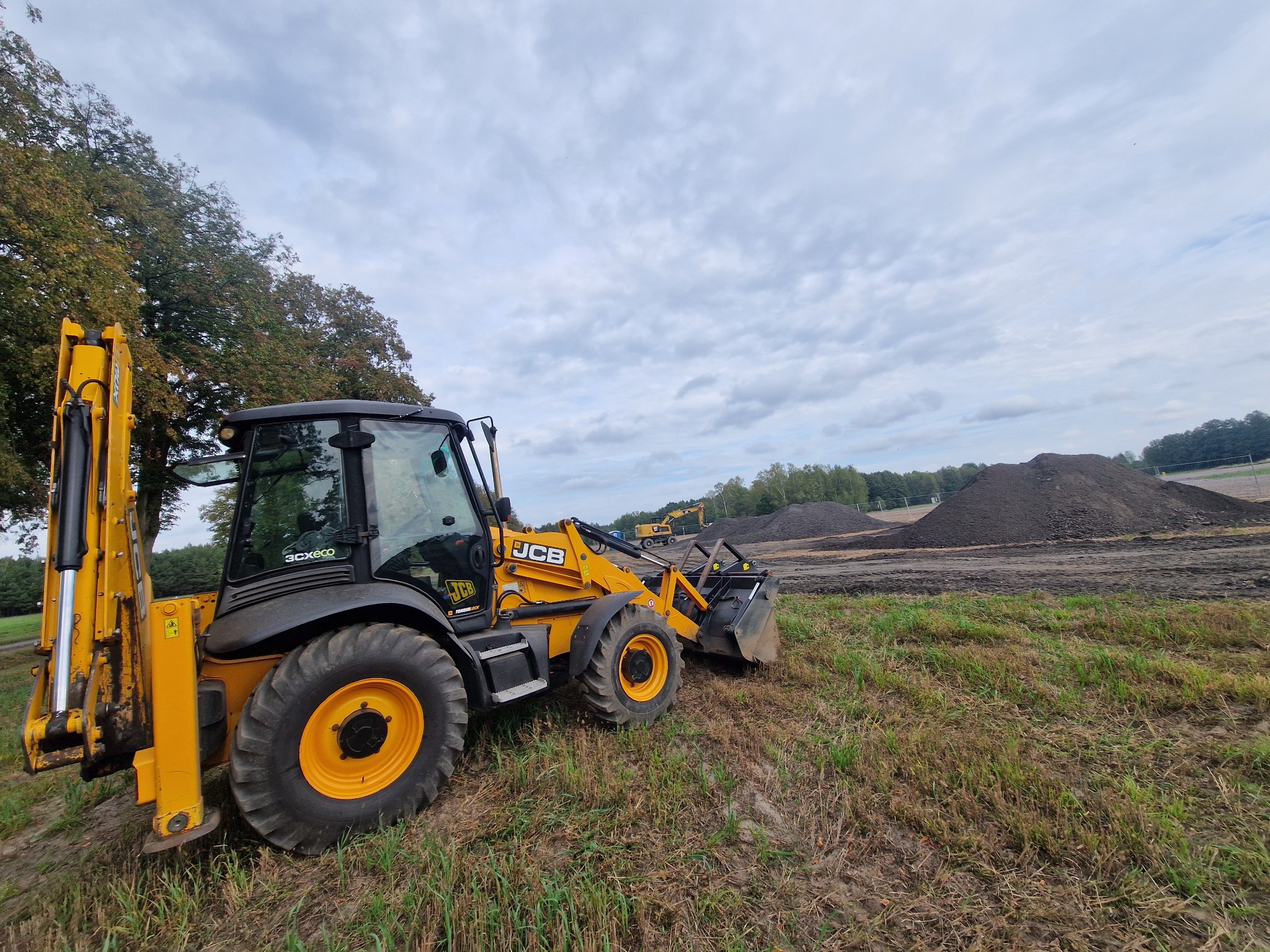 Żółto-czarna koparko-ładowarka JCB na polu z hałdami ziemi w tle, pod pochmurnym niebem. Widoczna druga koparka w oddali.