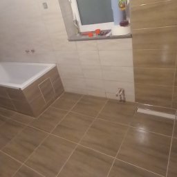 Usługi Ogólnobudowlane Michał Muzeja - Ukończona łazienka z brązowymi, podłużnymi płytkami na ścianach i podłodze, biała wanna zabudowana płytkami, okno z przyborami i butelką płynu do mycia, odpływ liniowy pod ścianą.