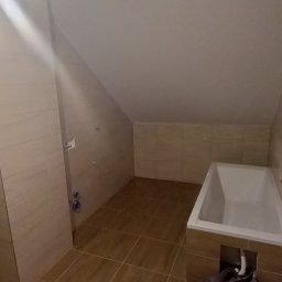 Usługi Ogólnobudowlane Michał Muzeja - Ukończona łazienka na poddaszu z brązowymi płytkami na podłodze i ścianach, biała wanna z obudową z płytek, narzędzia malarskie przykryte folią ochronną.