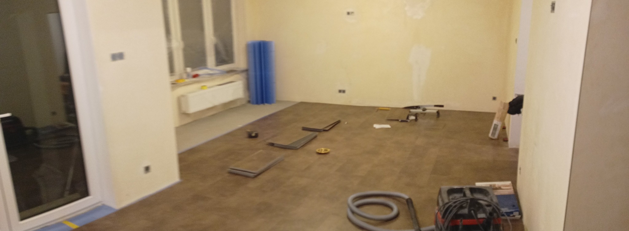 Wnętrze w trakcie remontu z brązową podłogą, narzędziami i odkurzaczem przemysłowym w rogu, ściany w kolorze ecru z widocznymi śladami po naprawach, okno z grzejnikiem i zwinięta mata izolacyjna.