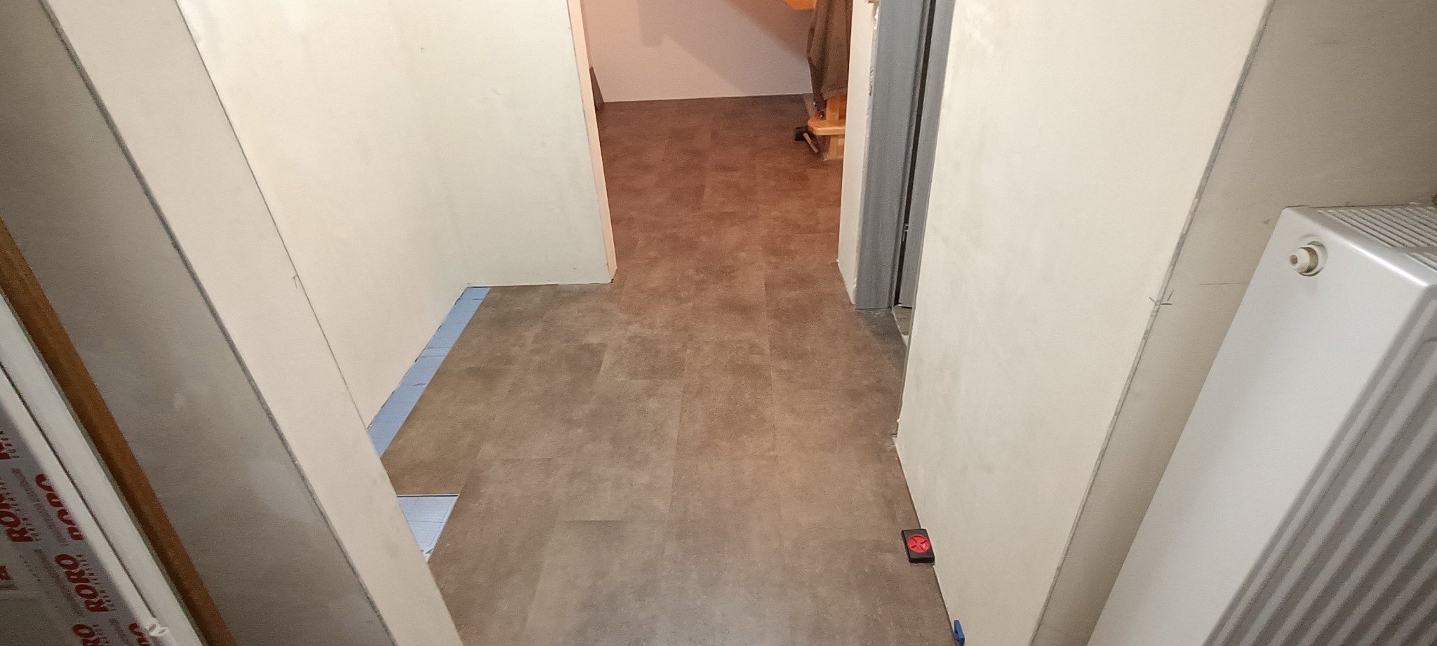 Korytarz w trakcie remontu: położona podłoga z paneli imitujących kamień, ściany przygotowane do malowania, widoczny grzejnik i fragment płyty przyściennej z nadrukiem.