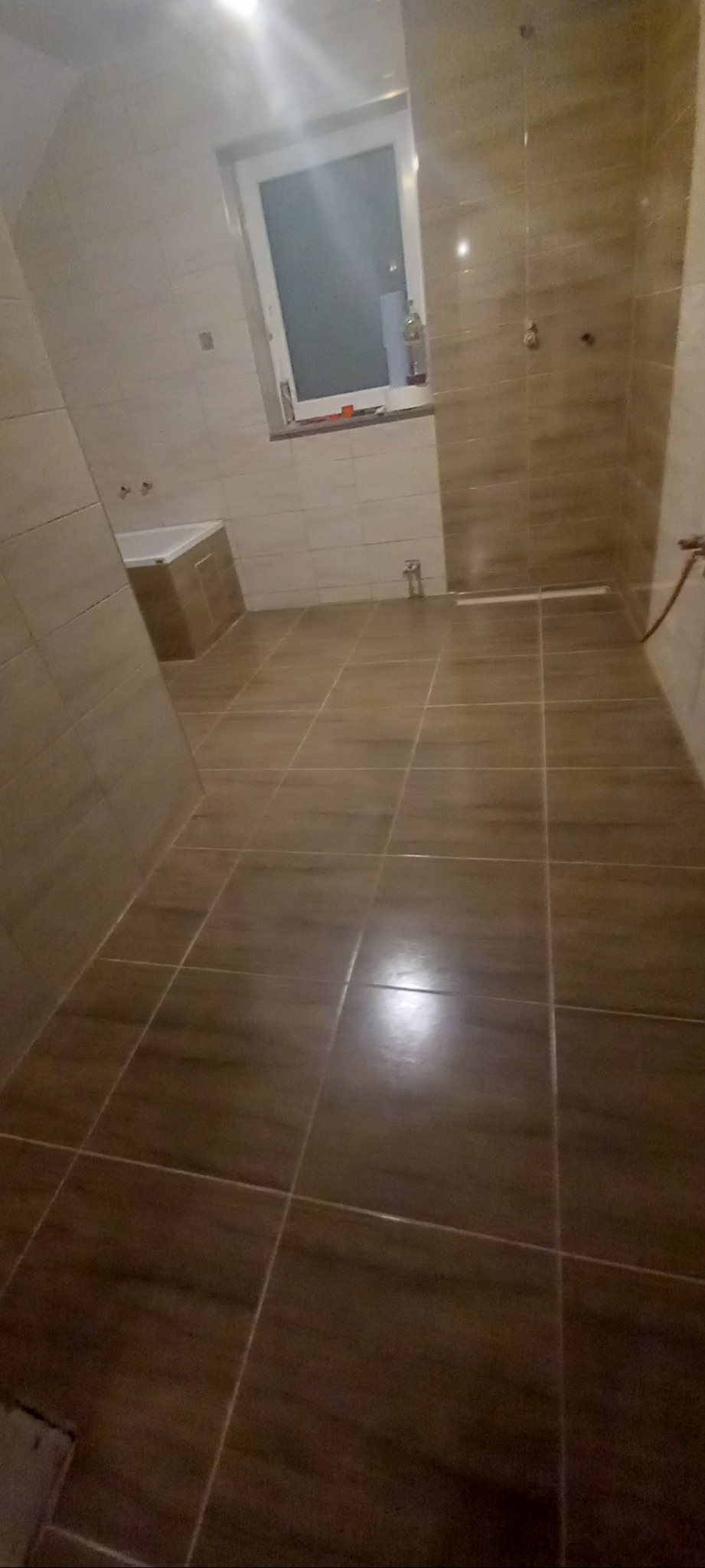 Wykończona łazienka z brązowymi, prostokątnymi płytkami na ścianach i podłodze, widoczna wanna przy ścianie i okno w tle.