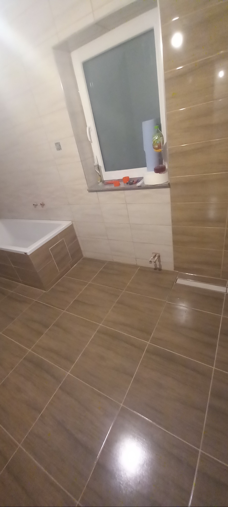 Ukończona łazienka z brązowymi, podłużnymi płytkami na ścianach i podłodze, biała wanna zabudowana płytkami, okno z przyborami i butelką płynu do mycia, odpływ liniowy pod ścianą.