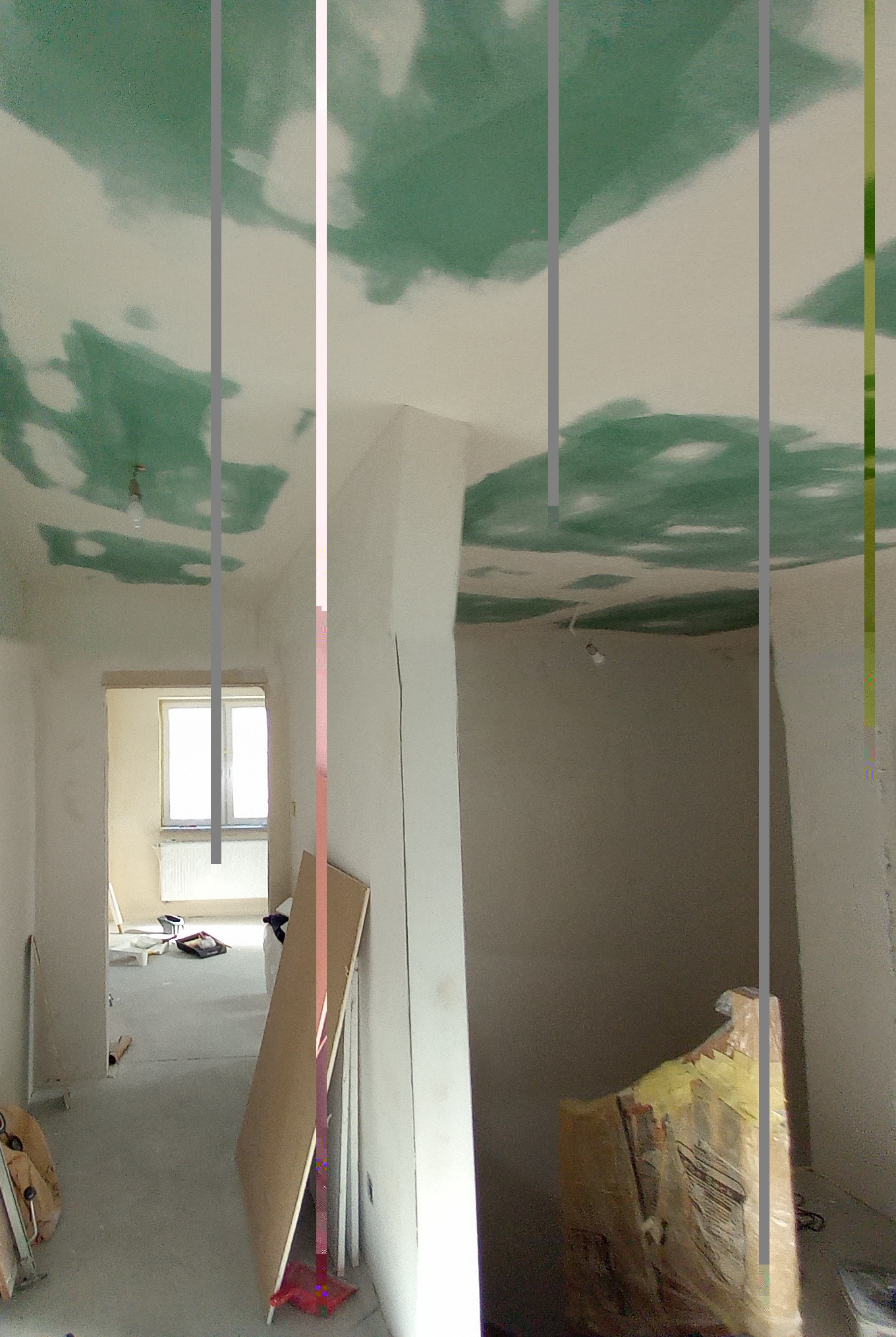 Wnętrze w trakcie prac wykończeniowych: sufit z widocznymi plamami po nałożeniu warstwy wyrównującej, ściany w stanie surowym, widoczne płyty przy ścianie, w tle okno z grzejnikiem.