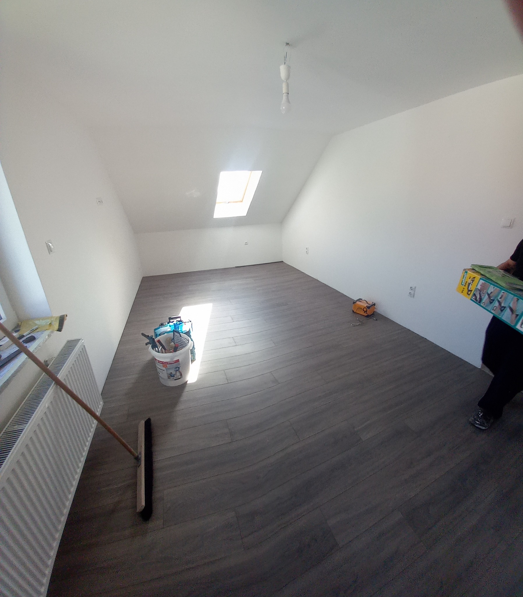 Poddasze z oknem dachowym i położoną podłogą z paneli imitujących drewno. Widoczne narzędzia i wiadro z akcesoriami malarskimi, oraz osoba wnosząca dodatkowe materiały.