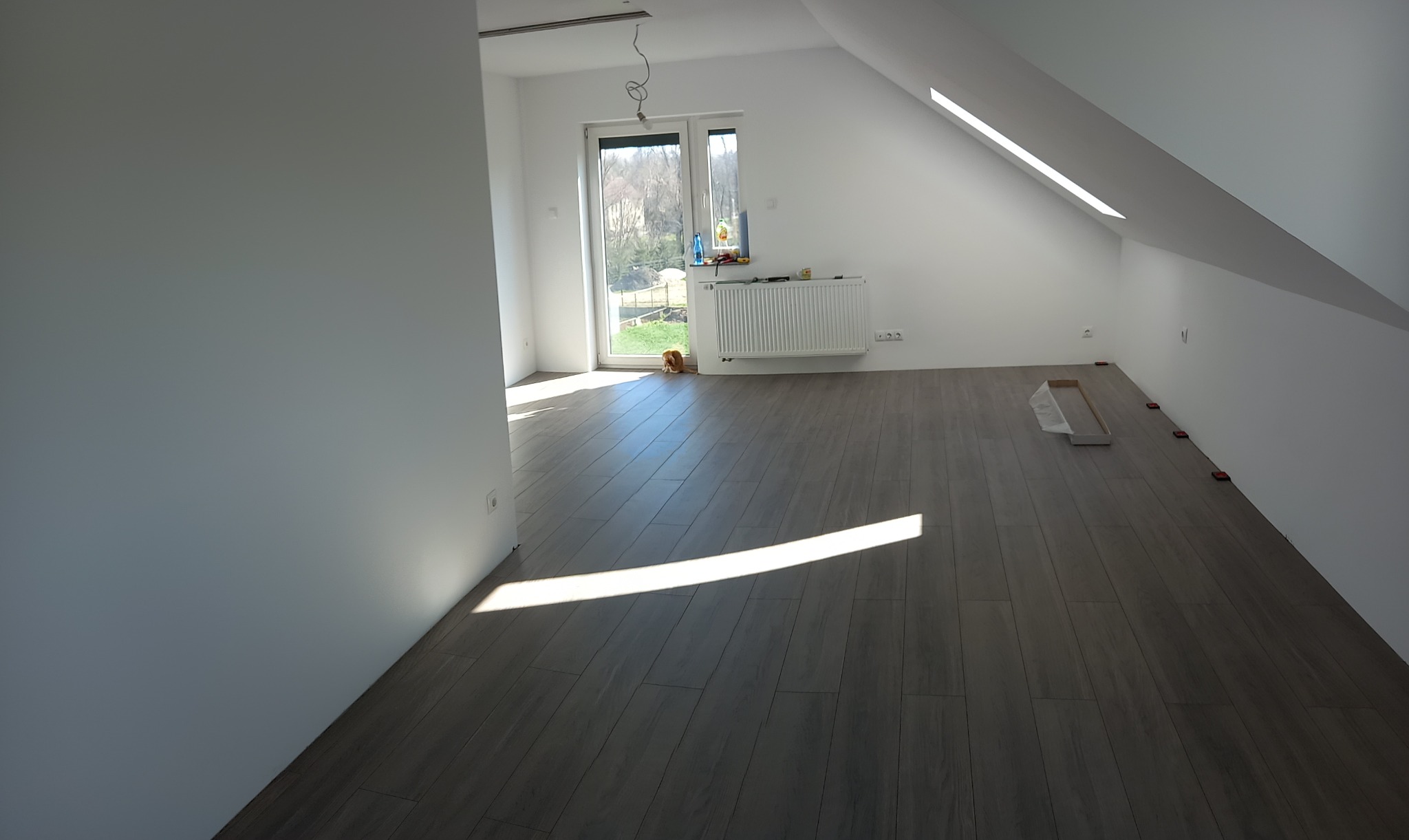 Poddasze z nową, ułożoną podłogą z paneli w odcieniach szarości, świeżo pomalowane na biało ściany, widoczne akcesoria montażowe, okno z widokiem na zewnątrz.