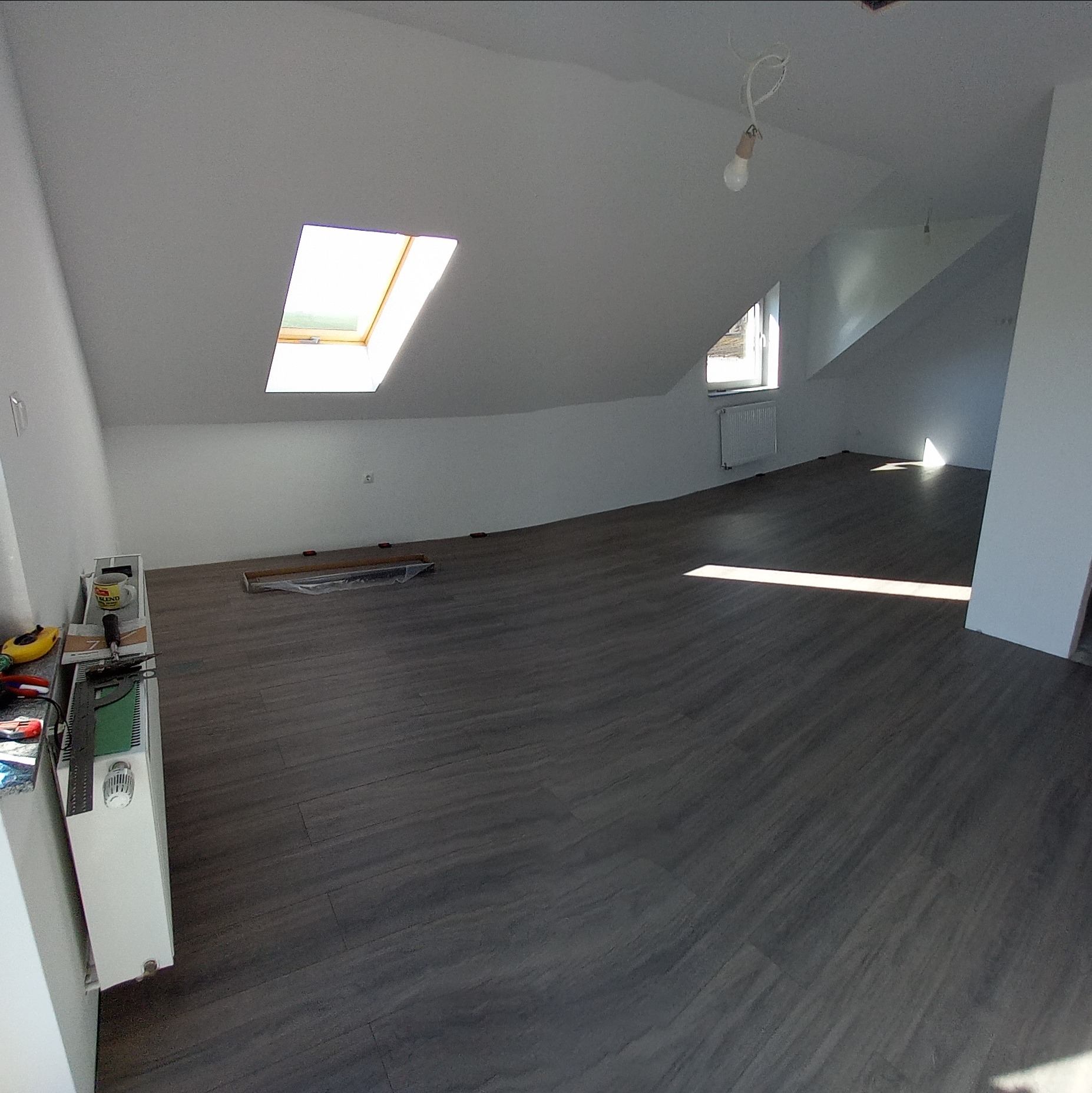 Pokój na poddaszu z nową, ciemną podłogą imitującą drewno, widoczne okno dachowe, grzejnik i narzędzia pracy na podłodze.