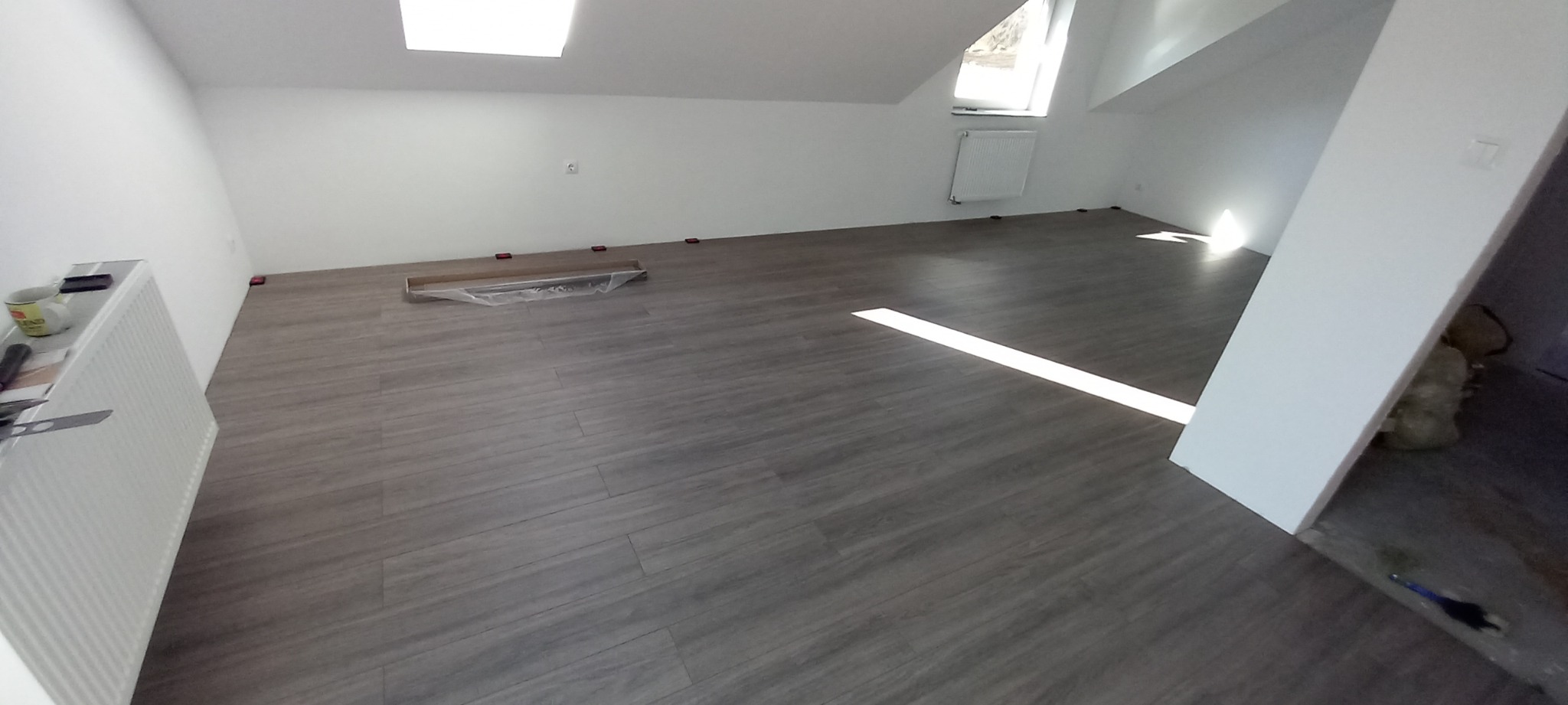 Układanie paneli podłogowych w nowo wykończonym, jasnym pokoju na poddaszu z oknem dachowym i grzejnikiem, widoczne elementy dystansowe i paczka paneli.