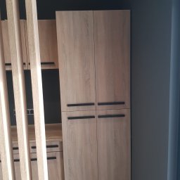 A.Meble Adrian Wojciechowski - Drewniana zabudowa meblowa z szafą dwudrzwiową i regałami, widoczna przez dekoracyjne, pionowe listwy, na tle ciemnoszarej ściany.