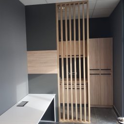 A.Meble Adrian Wojciechowski - Nowoczesne biuro z drewnianą ścianką działową, szafkami i białym biurkiem na tle szarych ścian.