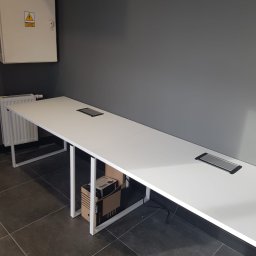 A.Meble Adrian Wojciechowski - Długi, biały stół konferencyjny z wbudowanymi gniazdami elektrycznymi, umieszczony w biurze z szarą ścianą i grzejnikiem.
