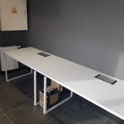 Długi, biały stół konferencyjny z wbudowanymi gniazdami elektrycznymi, umieszczony w biurze z szarą ścianą i grzejnikiem.