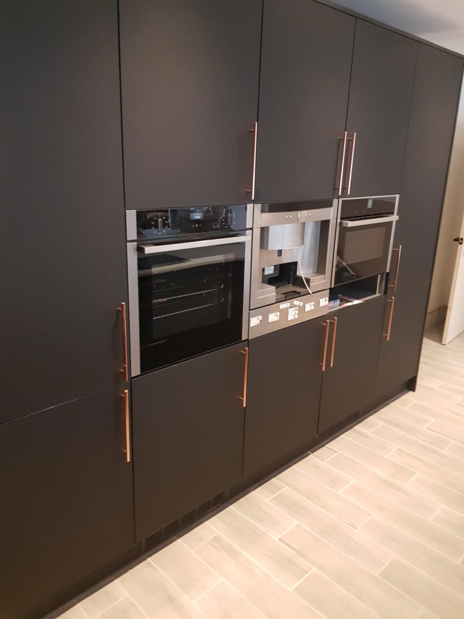Ciemna zabudowa kuchenna z wbudowanymi urządzeniami: piekarnik, ekspres do kawy i kuchenka mikrofalowa, z miedzianymi uchwytami, na tle jasnej podłogi z płytek.
