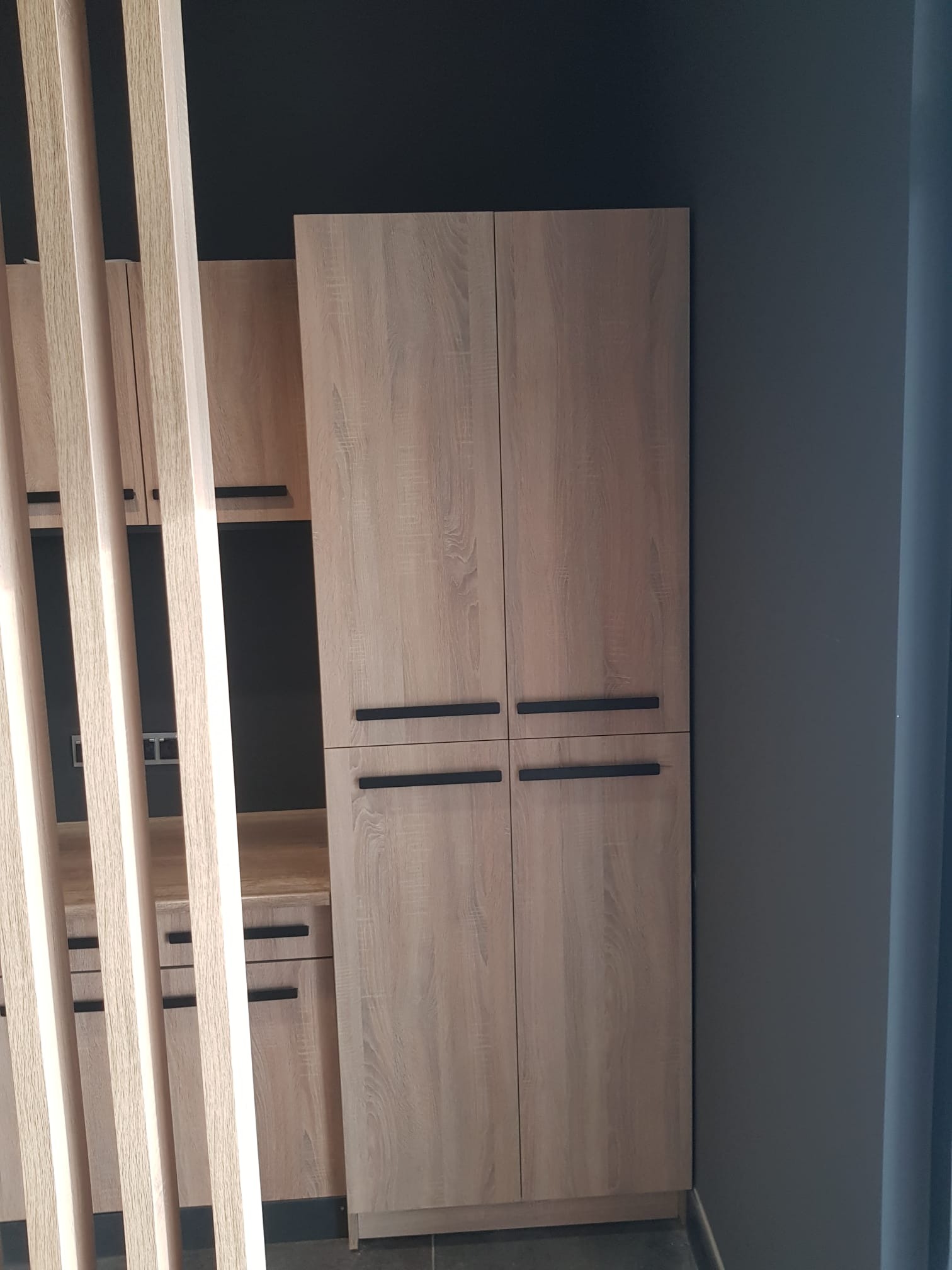 Drewniana zabudowa meblowa z szafą dwudrzwiową i regałami, widoczna przez dekoracyjne, pionowe listwy, na tle ciemnoszarej ściany.