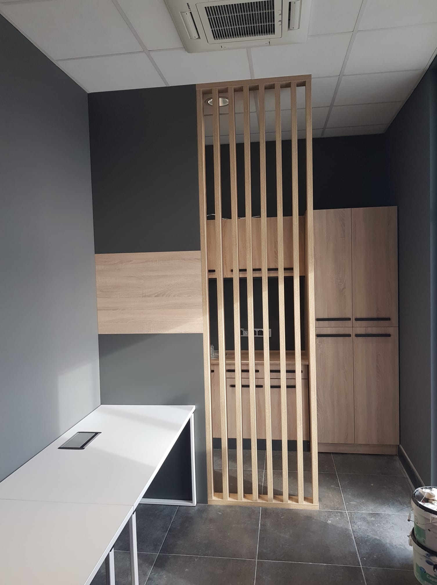 Nowoczesne biuro z drewnianą ścianką działową, szafkami i białym biurkiem na tle szarych ścian.