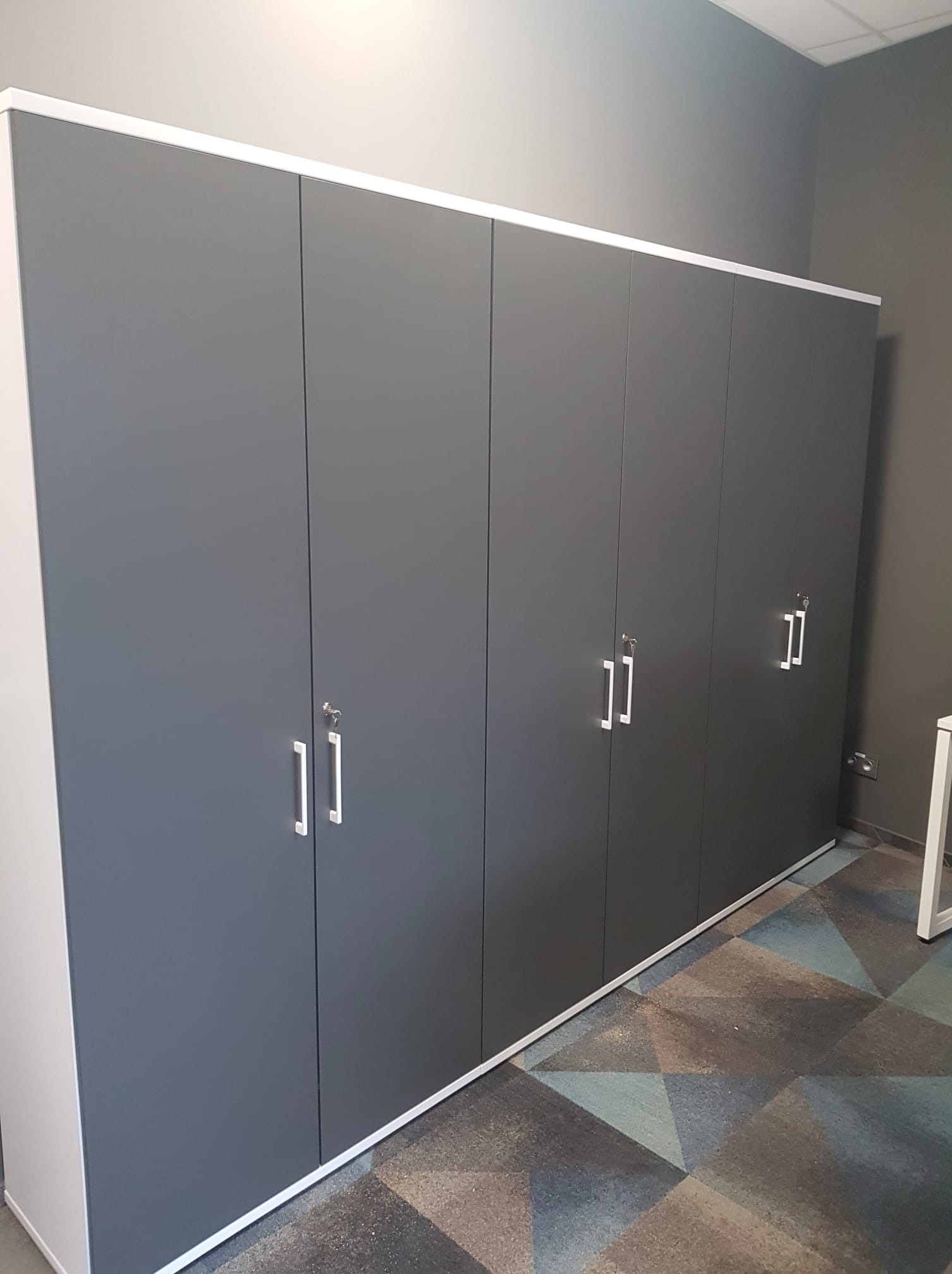 Szafa biurowa na całej ścianie z szarymi drzwiami i białymi obramowaniami, widoczne zamki w niektórych drzwiach, na podłodze wzorzysta wykładzina.