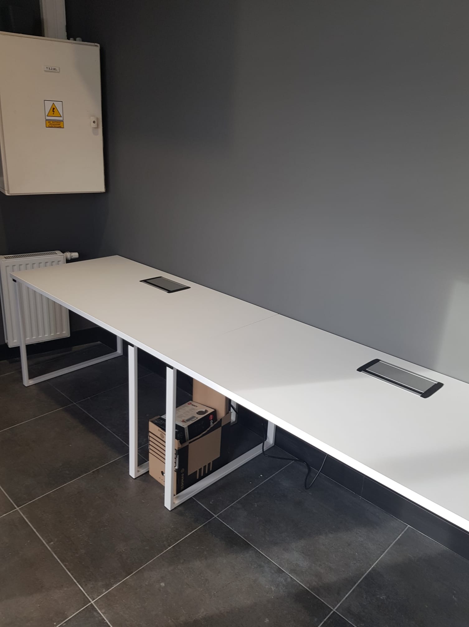 Długi, biały stół konferencyjny z wbudowanymi gniazdami elektrycznymi, umieszczony w biurze z szarą ścianą i grzejnikiem.