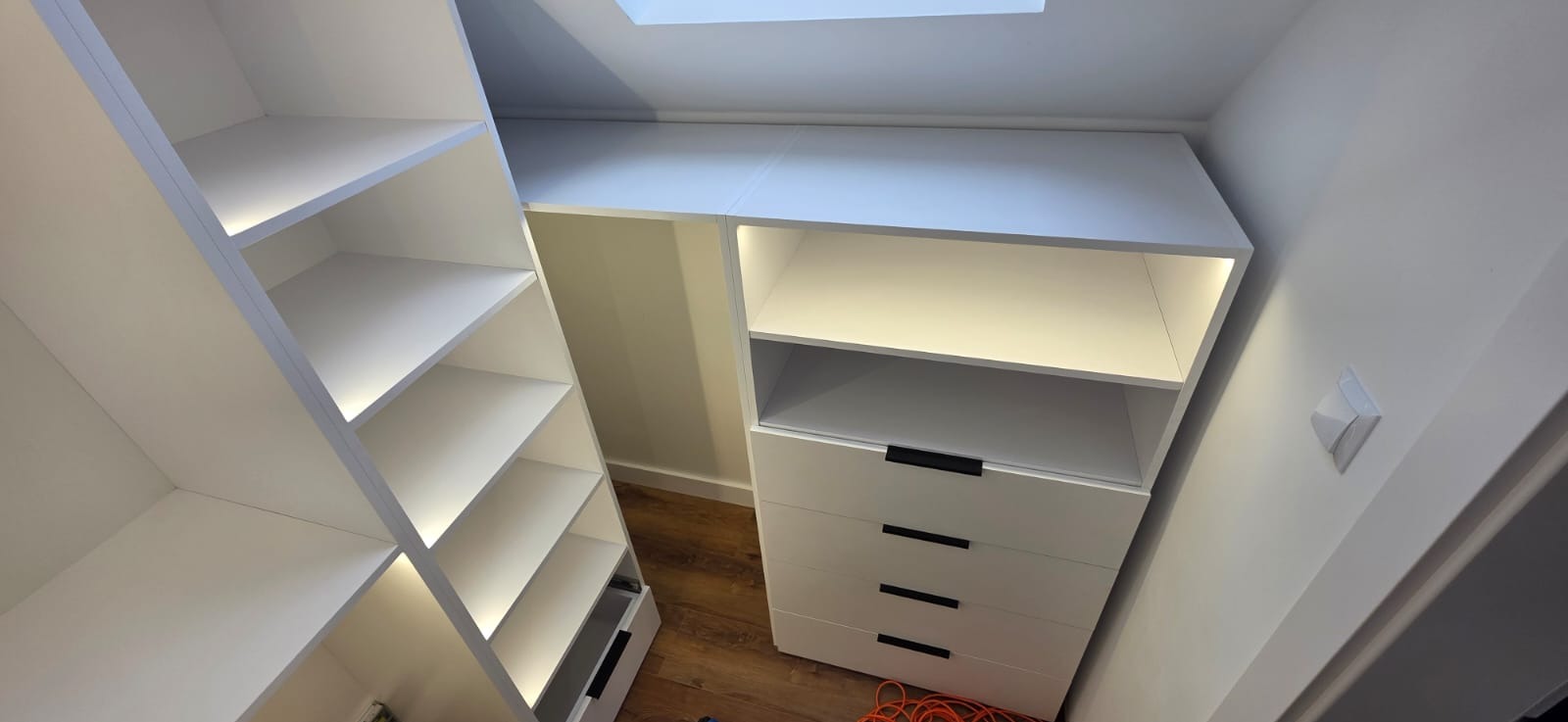 Biała zabudowa wnęki ze skośnym sufitem, z półkami oświetlonymi punktowo i komodą z czterema szufladami z czarnymi uchwytami, widok od dołu.