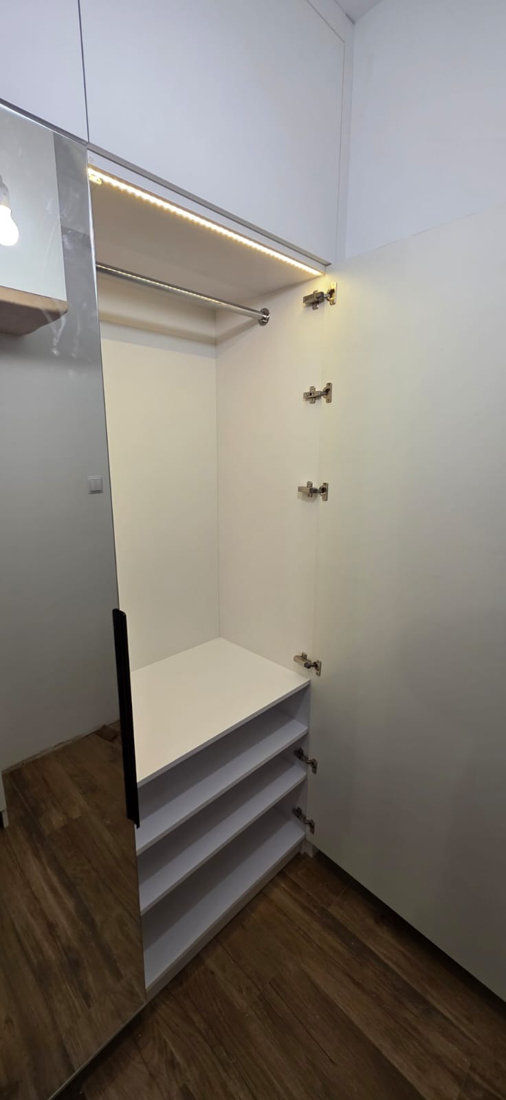 Wnętrze białej szafy z drzwiami uchylnymi, oświetleniem LED i półkami, widoczne zawiasy i lustrzane drzwi po lewej stronie.