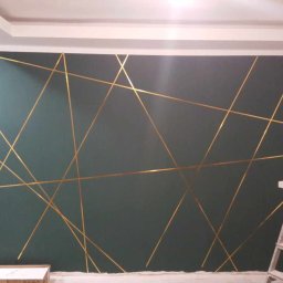 Ekspert Pięknego Wnętrza - Ciemnozielona ściana z dekoracyjnymi, złotymi liniami tworzącymi geometryczny wzór, widoczna drabina i materiały budowlane, drzwi w tle.