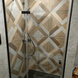 Kabina prysznicowa z czarnymi okuciami i wzorzystymi płytkami w odcieniach drewna i szarości, widok z góry na brodzik i częściowo widoczne narzędzia w tle.