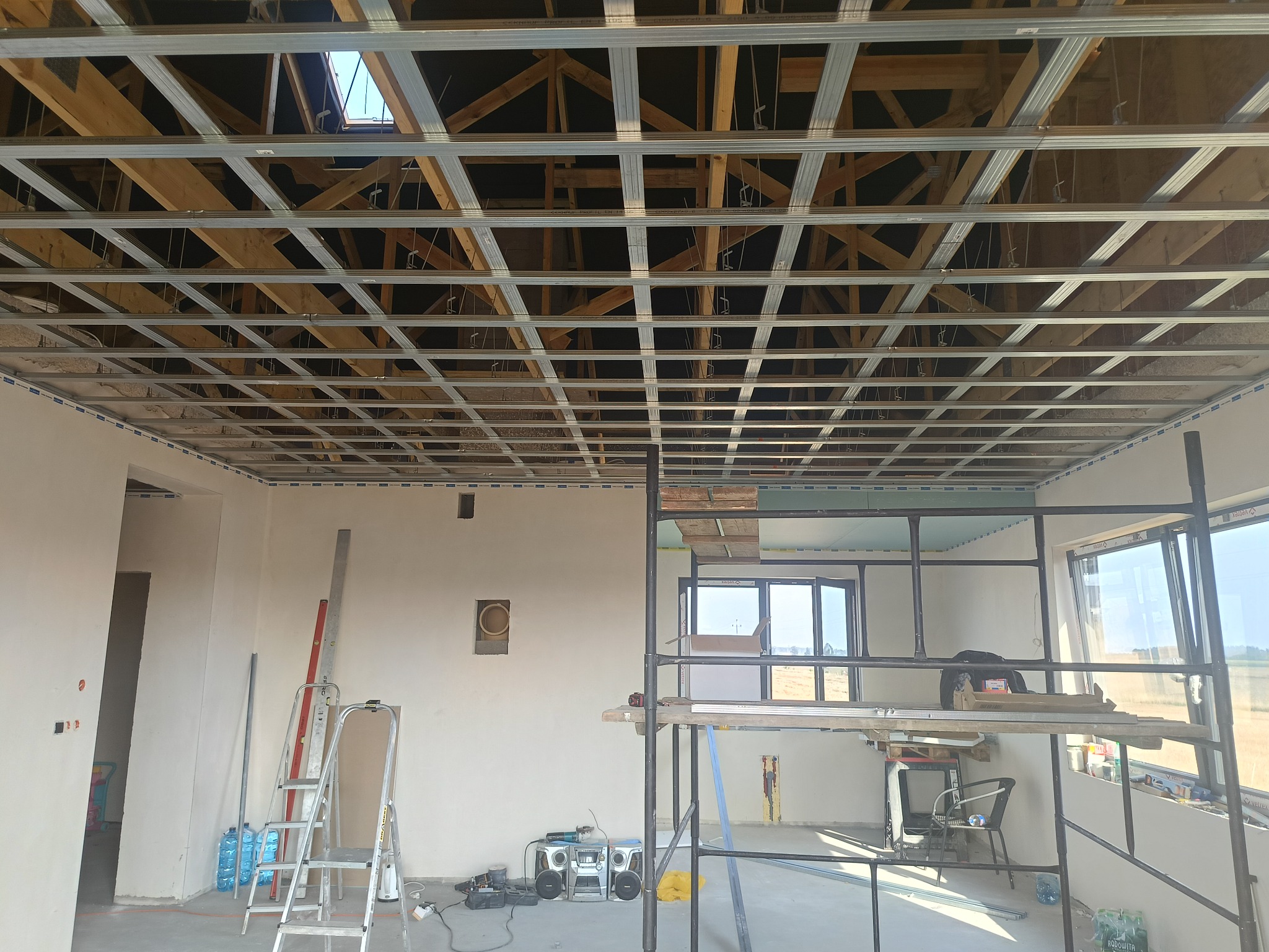 Wnętrze w trakcie prac wykończeniowych: widoczna metalowa konstrukcja podwieszanego sufitu z drewnianą więźbą dachową powyżej, ściany w stanie surowym, drabina, rusztowanie, narzędzia oraz okna...