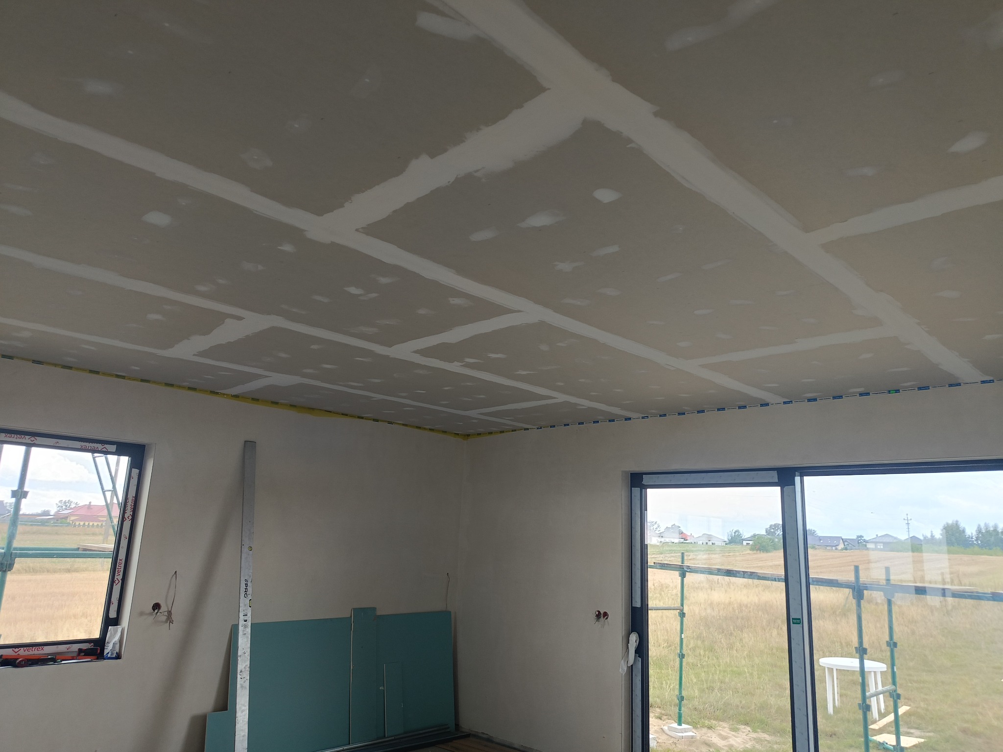 Sufit z płyt gipsowo-kartonowych po szpachlowaniu łączeń, widoczne taśmy wzmacniające, w tle okno z widokiem na zewnątrz i pionowa poziomica oparta o ścianę.