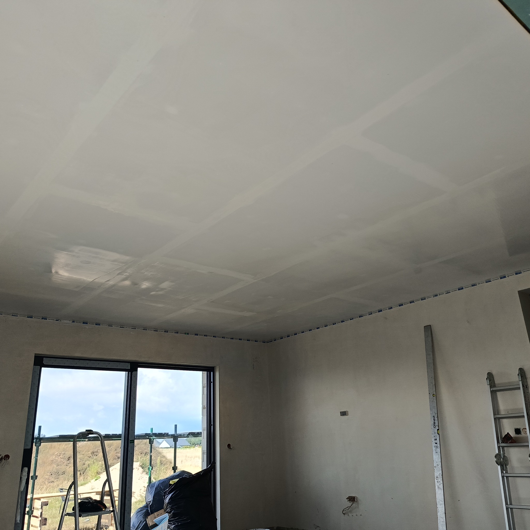 Wnętrze w trakcie prac wykończeniowych: wyszpachlowany sufit z widocznymi łączeniami płyt gipsowo-kartonowych, ściany przygotowane do malowania, okno z widokiem na zewnątrz, drabina oparta o ścianę.