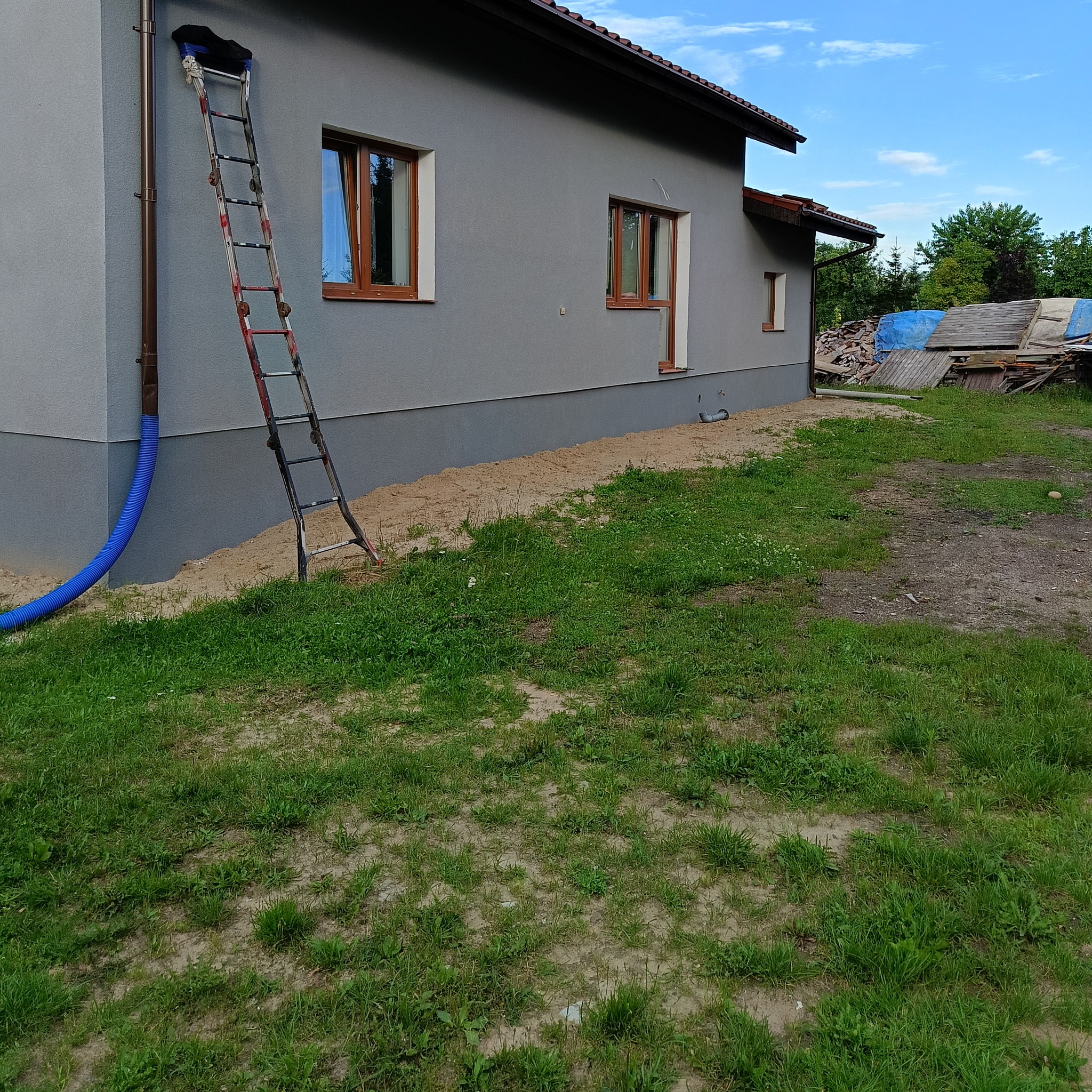 Szary dom jednorodzinny z brązowymi oknami, z zakończonym ociepleniem fundamentów, z niebieskim wężem odprowadzającym wodę i drabiną opartą o rynnę, na trawniku