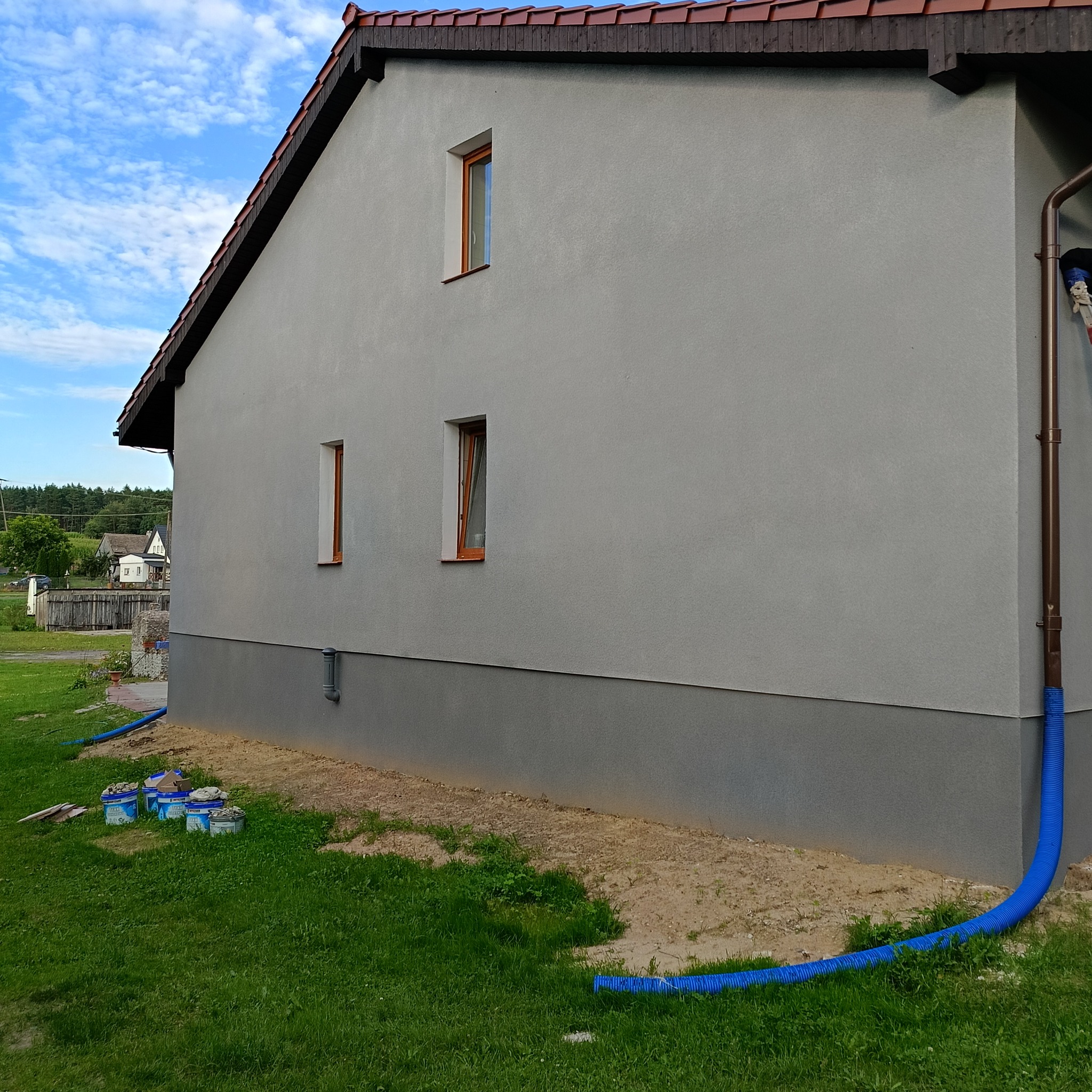 Świeżo otynkowana ściana zewnętrzna domu z widocznymi oknami z drewnianą ramą, rynną, niebieskim wężem odprowadzającym wodę i wiaderkami z zaprawą na trawie.