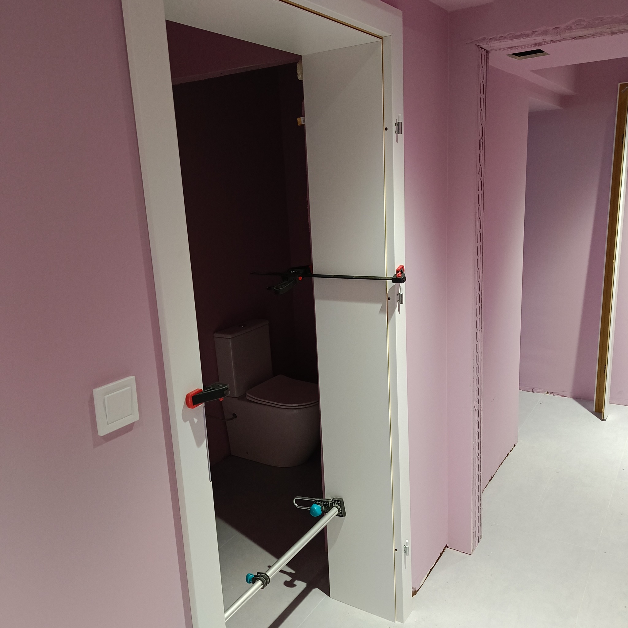 Białe drzwi wewnętrzne, zamocowane rozpórkami montażowymi w ościeżnicy, widok na toaletę z muszlą klozetową w pomieszczeniu z różowymi ścianami.