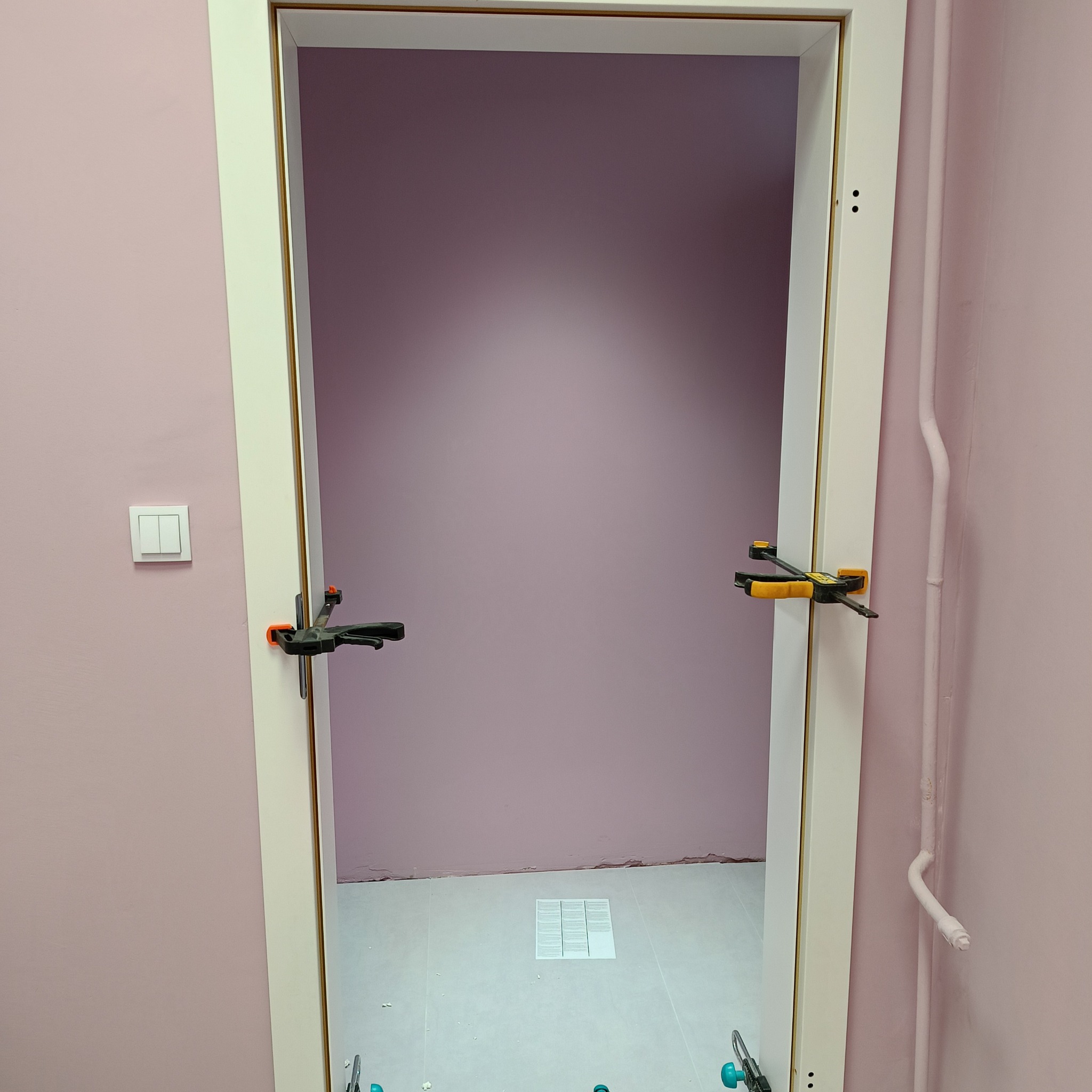 Biała ościeżnica drzwiowa mocowana za pomocą dwóch ścisków stolarskich w pomieszczeniu z różowymi ścianami i jasnymi płytkami podłogowymi.