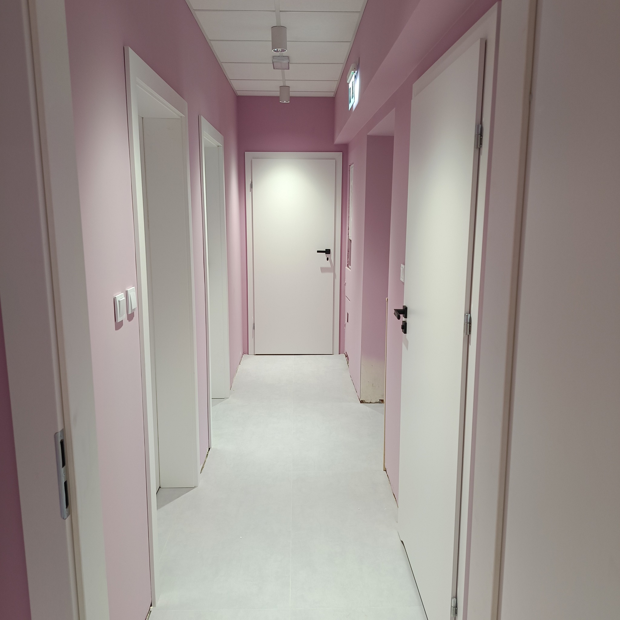Korytarz po remoncie z różowymi ścianami, białymi drzwiami z czarnymi klamkami i jasną podłogą, widoczne oświetlenie sufitowe.