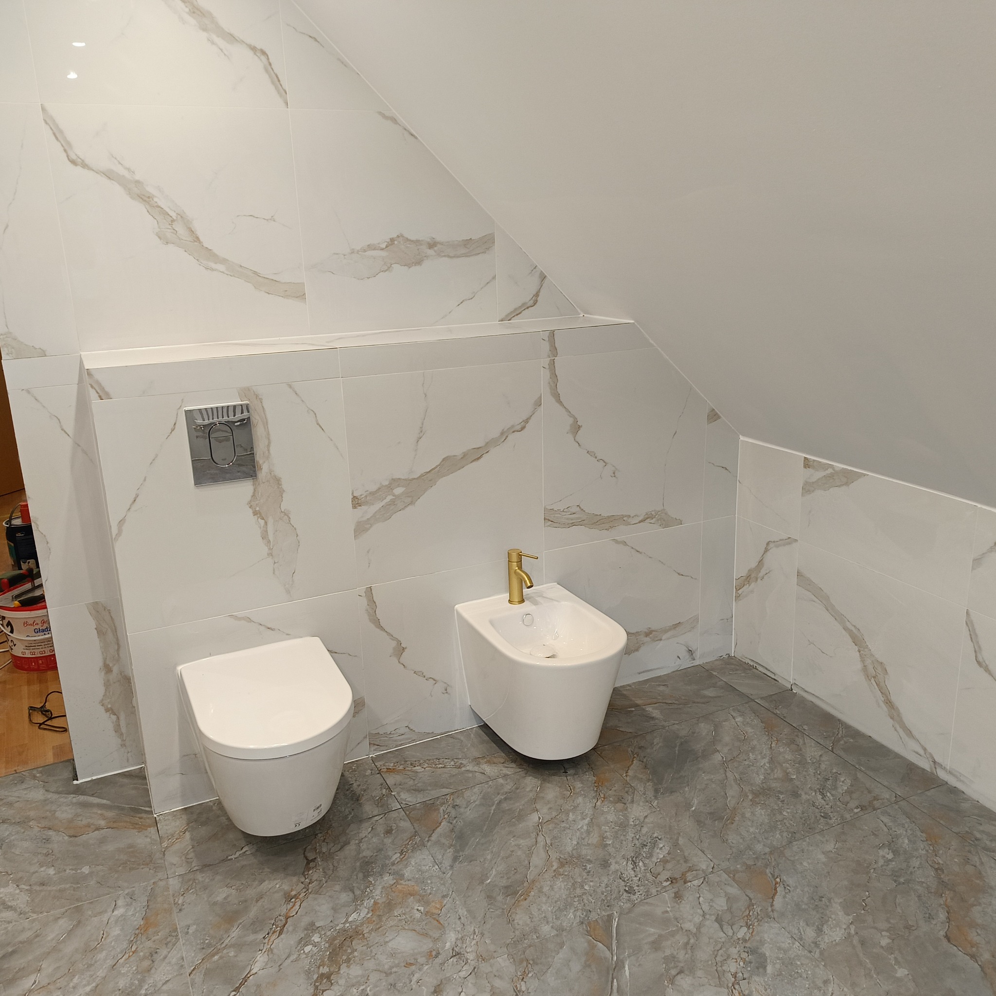 Nowoczesna łazienka na poddaszu z toaletą i bidetem, wykończona białymi płytkami imitującymi marmur z delikatnymi złotymi żyłkami, oraz szarą podłogą z motywem marmuru. Złota bateria przy bidecie...