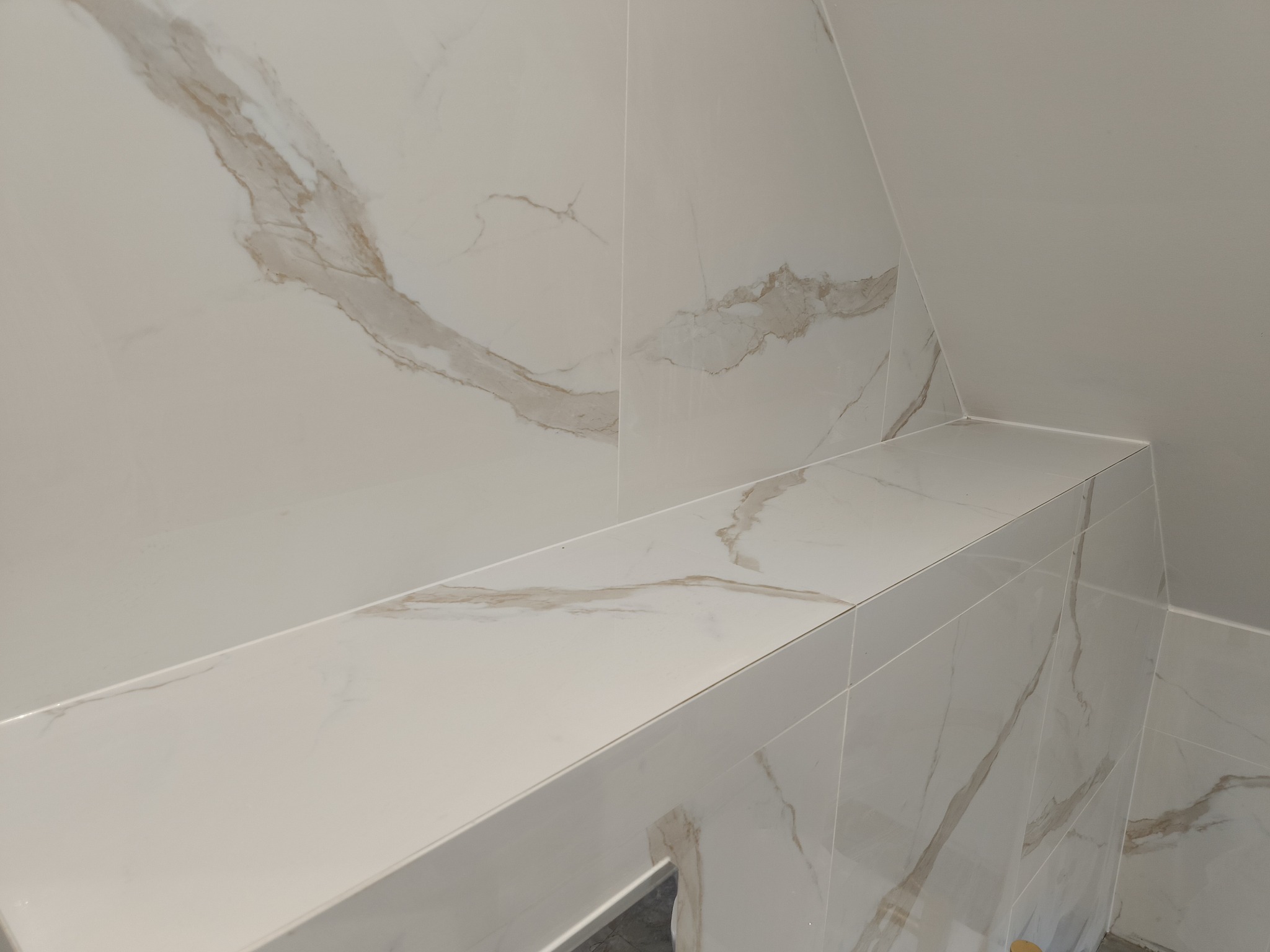 Półka z płytek imitujących marmur w narożniku łazienki ze skośnym sufitem, widoczne fugi i wzór żyłkowania.