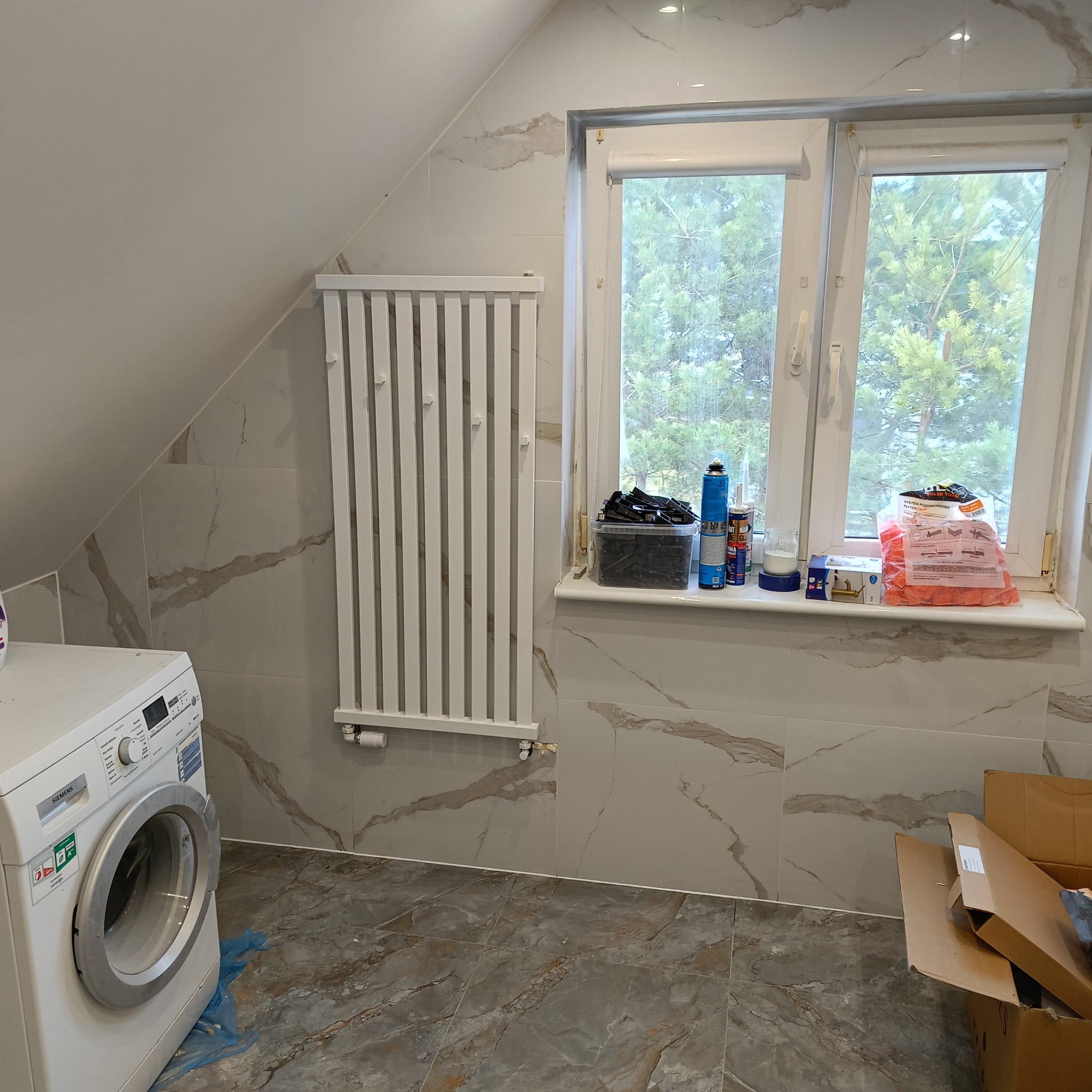 Ukończona łazienka na poddaszu z marmurowymi płytkami, grzejnikiem drabinkowym, pralką Siemens i materiałami budowlanymi na parapecie okna.
