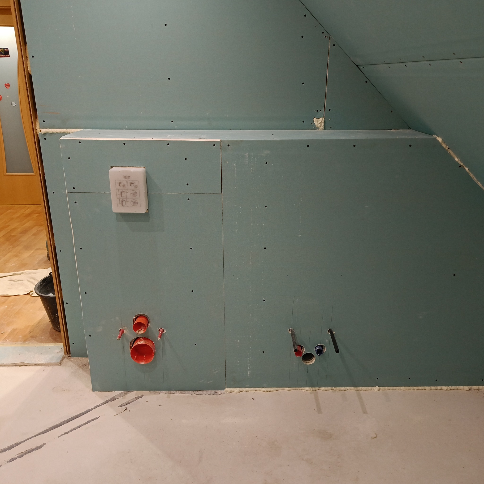 Ściana z płyt gipsowo-kartonowych w trakcie montażu w łazience, widoczne otwory na instalacje wodno-kanalizacyjne, puszka podtynkowego panelu sterowania armaturą Grohe, ślady po cięciu w betonie...