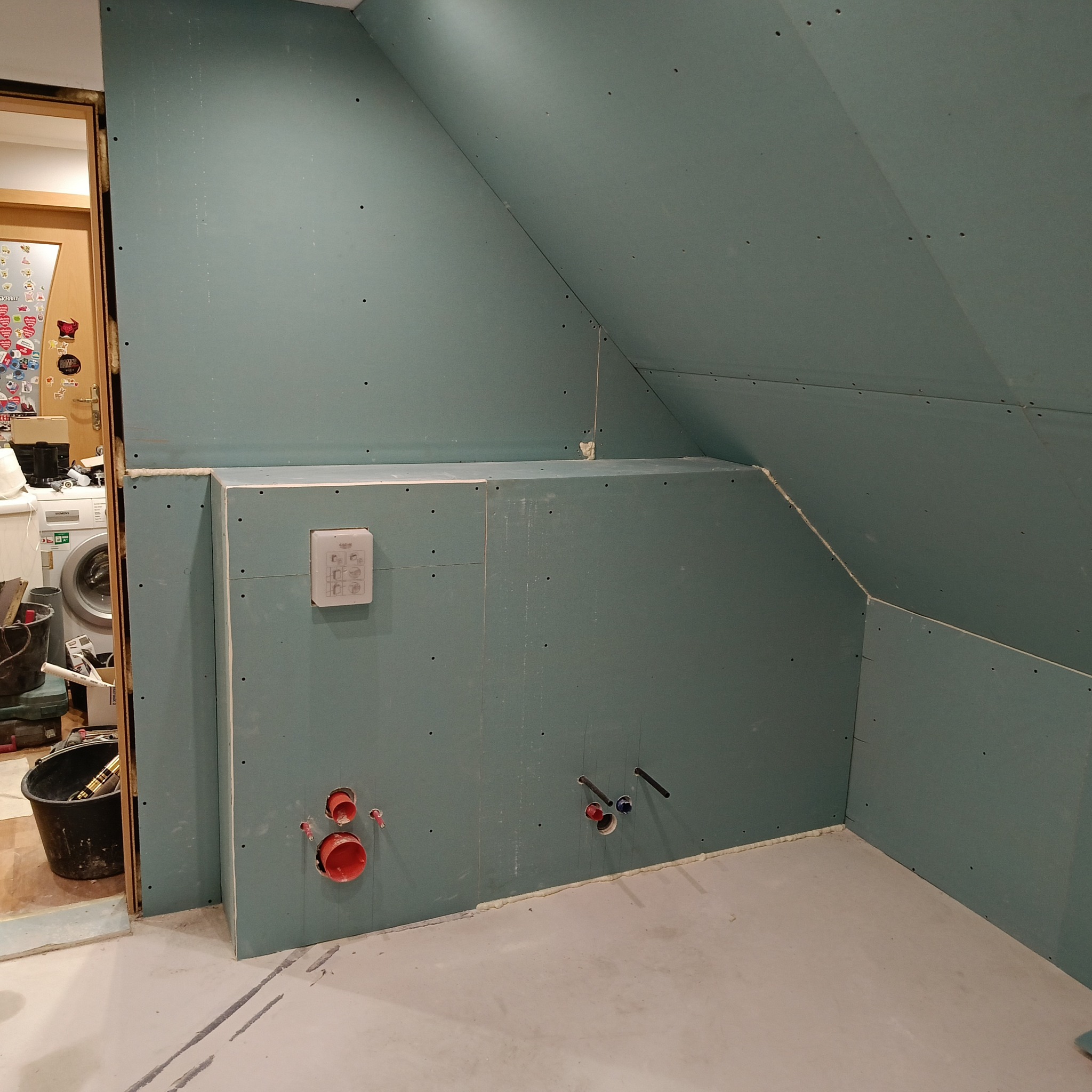 Ściany z płyt gipsowo-kartonowych w łazience na poddaszu, widoczne instalacje wodne, puszka podtynkową, w tle pralka i narzędzia.