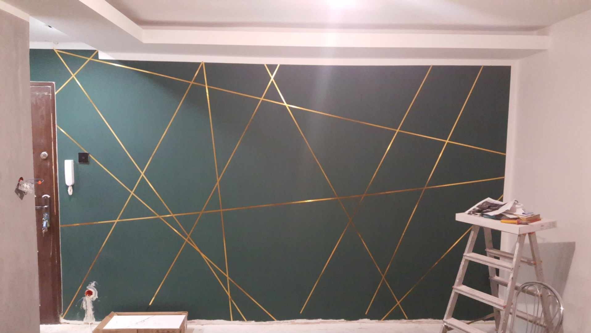 Ciemnozielona ściana z dekoracyjnymi, złotymi liniami tworzącymi geometryczny wzór, widoczna drabina i materiały budowlane, drzwi w tle.