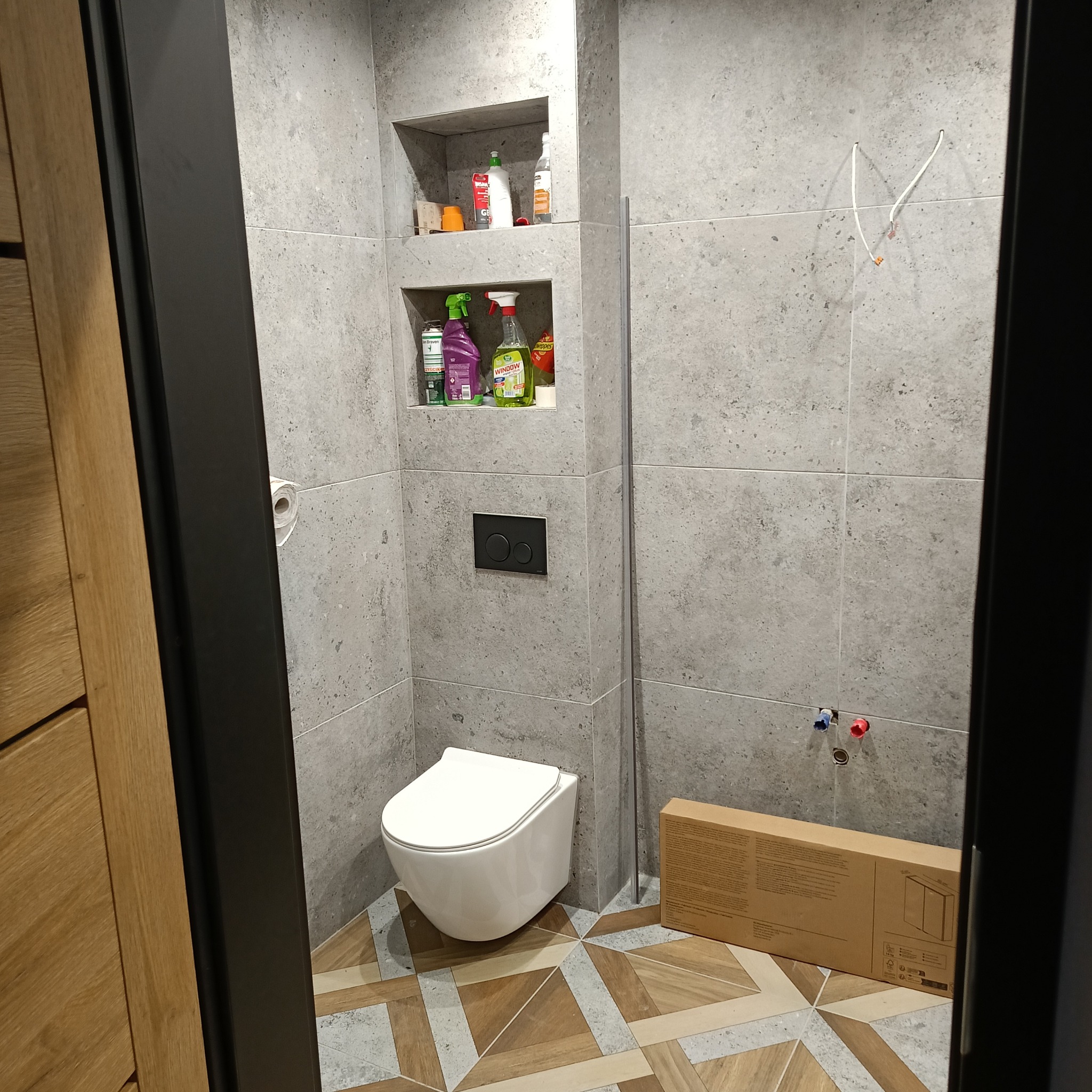 Nowoczesna łazienka z wiszącą toaletą, szarą okładziną ścienną z wnękami na akcesoria, dekoracyjną podłogą i widocznymi przyłączami wodnymi przed montażem armatury.