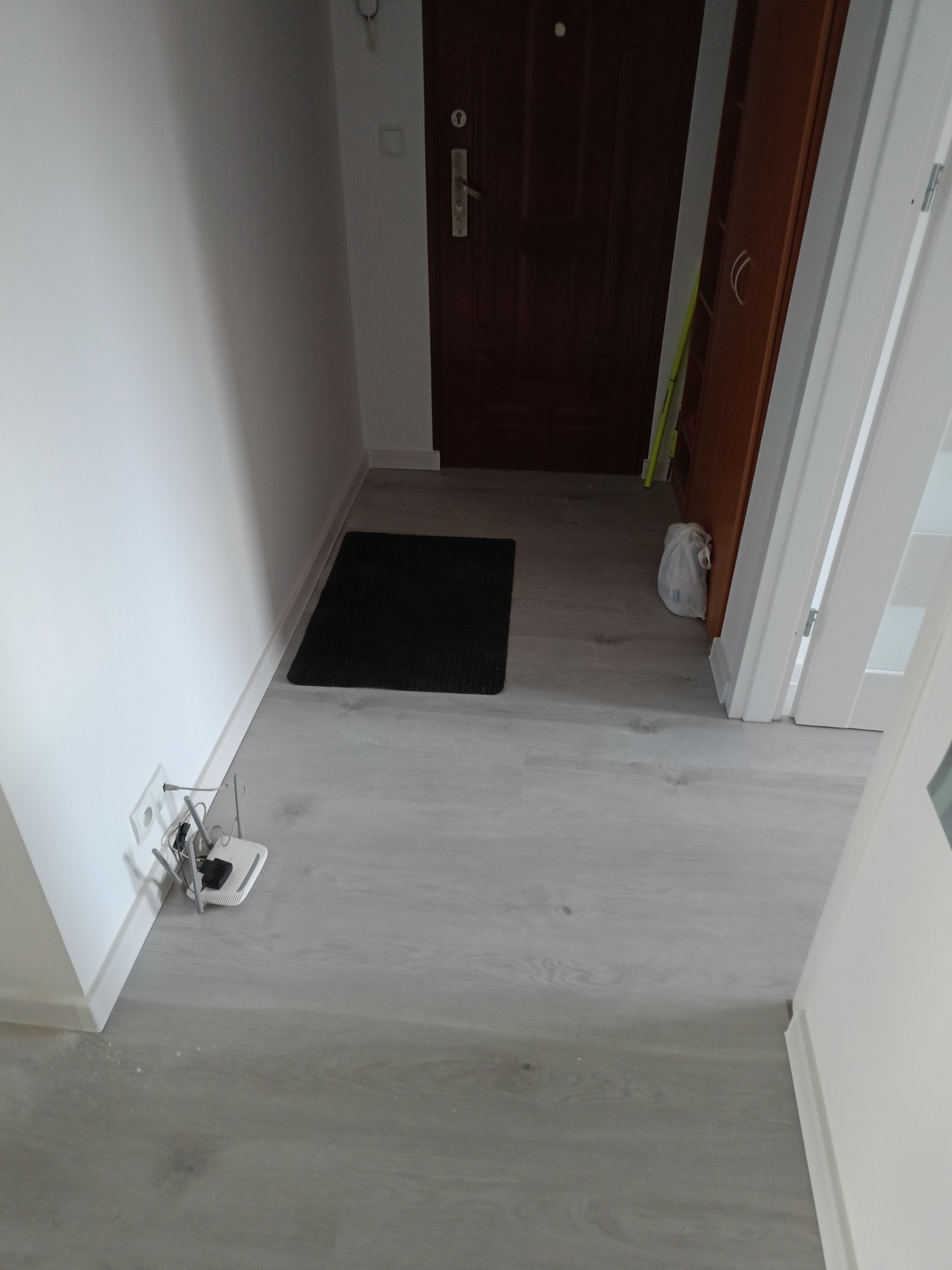 Korytarz z szarą podłogą imitującą drewno, ciemnymi drzwiami wejściowymi, brązową szafką i czarnym dywanikiem przy progu; widoczny router przy ścianie.